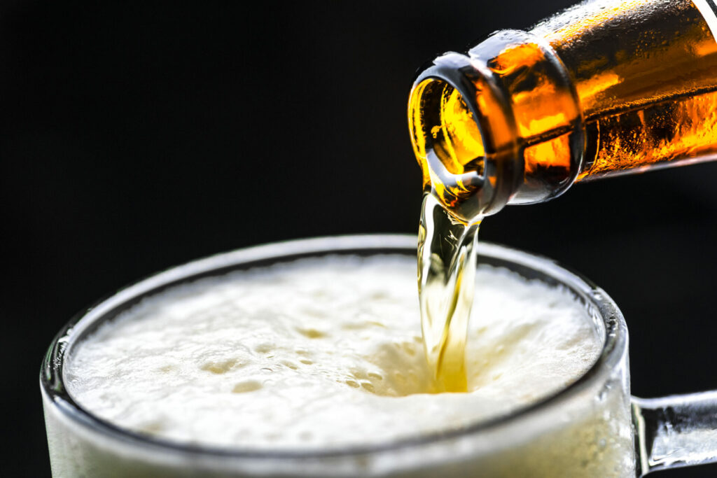 Dettaglio di una bottiglia di birra che verso schiuma densa in un boccale trasparente su sfondo scuro - attualita.it Dettaglio di una bottiglia di birra che verso schiuma densa in un boccale trasparente su sfondo scuro