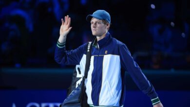 Tennis player in a blue cap and jacket waves to the crowd, carrying a bag on a dark blue court.