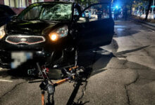 Nighttime street scene showing a black SUV with a shattered windshield and open front door after a collision; a bike-like device lies on the cracked pavement in front of the vehicle, with emergency responders and blue lights in the background.