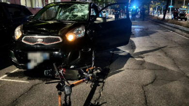 Nighttime street scene showing a black SUV with a shattered windshield and open front door after a collision; a bike-like device lies on the cracked pavement in front of the vehicle, with emergency responders and blue lights in the background.